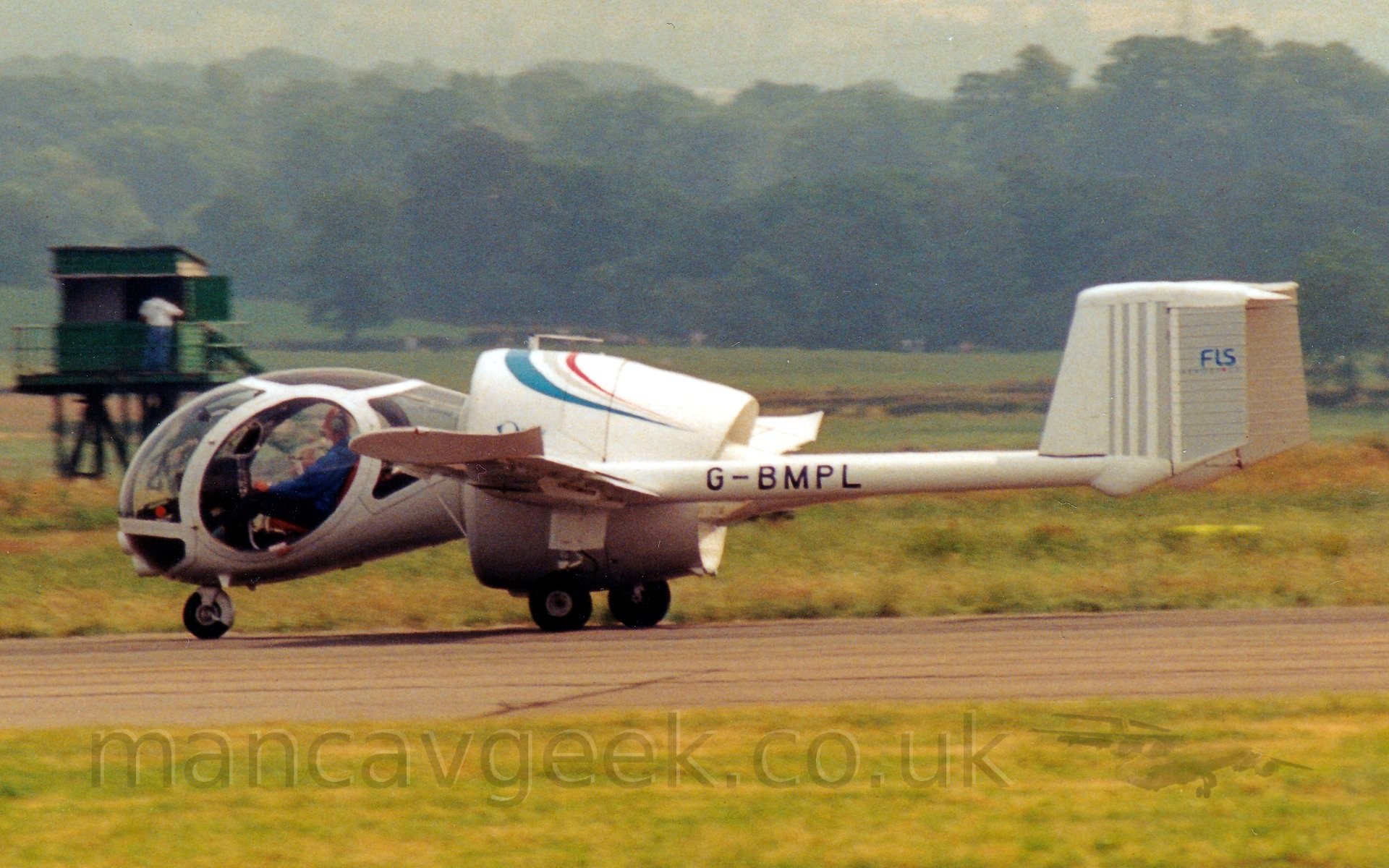 Side view of a single-engined light aircraft taxiing from right to left along a black runway.
The plane is mostly white, with an engine inside a large shroud in the middle, looking much like a jet engine, with wings sticking out from the side, long twin boom extending from the rear of the wings carrying the tail.
The black registration "G-BMPL" is shown on these booms.
The cockpit is a complex, heavily glazed ovoid, almost a classic rear-drop shape, sticking out from the front of the engine shroud, and giving the plane an almost insectoid appearance.
Lush green grass lines the runway in the foreground, with more grass in the background leading up to trees in the distance, under hazy grey skies.