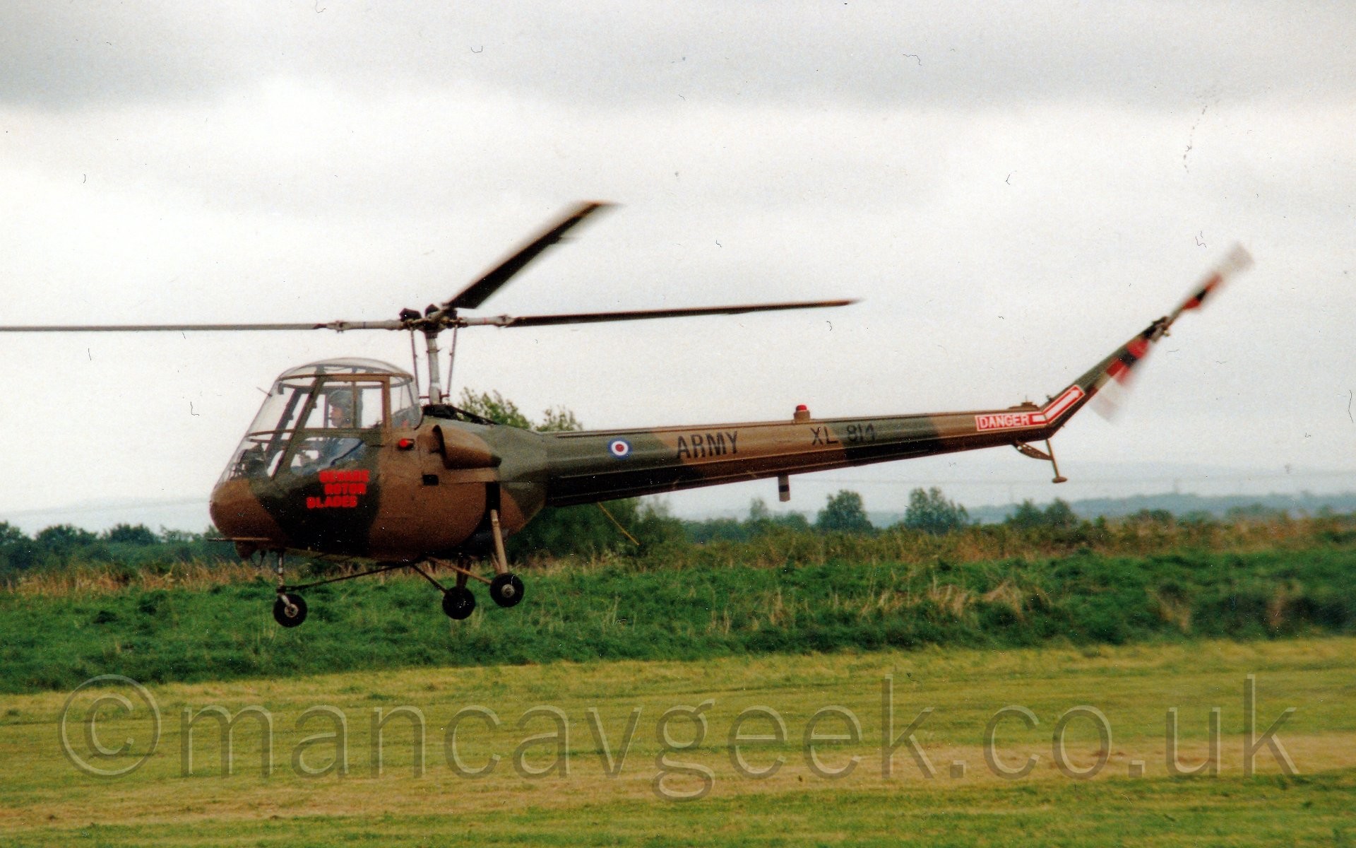 Side view of a small, rather ancient-looking helicopter flying from right to left above a grass airfield.
The helicopter is covered in an olive green and brown camouflage scheme, with red "Beware rotor blades" text below the upturned-bucket cockpit.
There is black "Army" titles and the serial "XL814" on the tail boom, as well as a red block with white text "Danger" and a white arrow pointing to the spinning tail rotor.
It is flying just a few metres above a grass field, with bushes and trees in the distance, under flat grey sky.